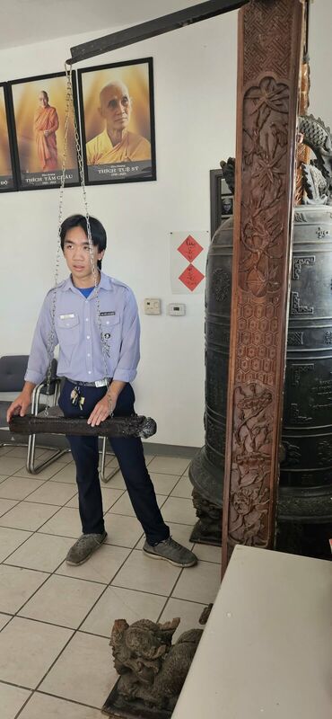 Huynh Trưởng tập sự cầm dùi chuông bên đại hồng chung, phía sau là chân dung chư Tôn Đức